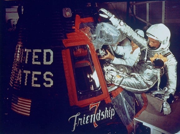 Hombre con traje plateado retorciéndose con los pies por delante en una pequeña cápsula espacial.