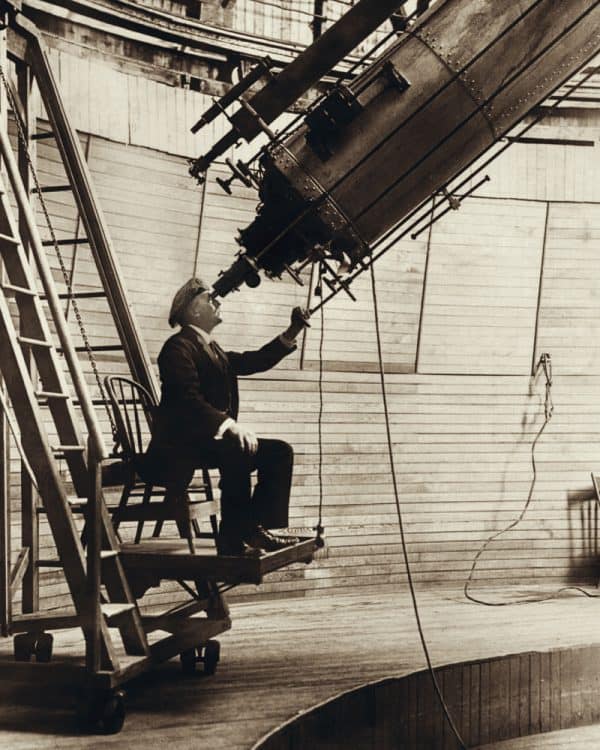 Hombre con ropa de principios del siglo XX, sentado en el ocular de un gran telescopio.