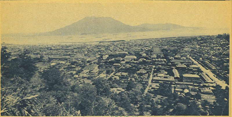 Foto antigua de una gran ciudad japonesa de estilo antiguo cubierta de cenizas, con un volcán visible en la distancia.